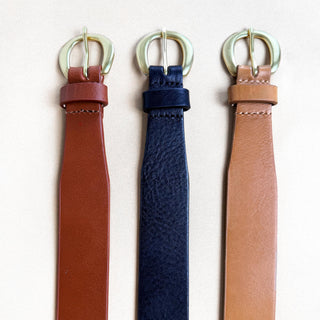 Three leather belts with round metal buckles, shown vertically side by side on a light beige background. The belts are in brown, navy blue, and tan, with the buckles appearing gold-toned.